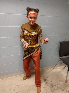 a person with makeup on their face standing next to a chair and wearing an orange outfit