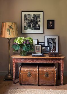 a wooden table with flowers and pictures on it next to a lamp in a living room