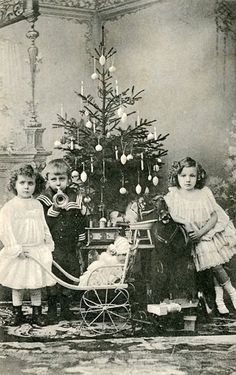 three children standing next to a christmas tree in front of a small sleigh