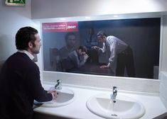 a man standing in front of a bathroom mirror next to two sinks and a large poster on the wall