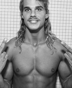 a man with long hair and no shirt standing in front of a tiled wall holding his hands up to his chest