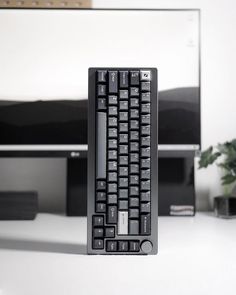 a computer keyboard sitting on top of a white desk next to a monitor and plant