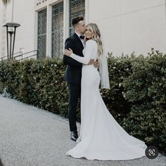 a bride and groom embracing in front of a white building with greenery on the sidewalk