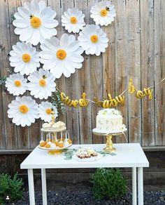 a white table topped with a cake next to a wooden fence covered in paper flowers