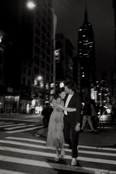 a man and woman crossing the street at night in new york city, with skyscrapers in the background