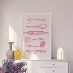 a pink kitchen art print on a white cabinet next to purple flowers and vases