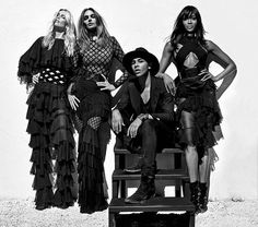 three women and one man sitting on a bench in front of a white wall wearing black clothing