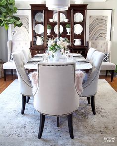 a dining room table set for four with white chairs and china cabinet in the background