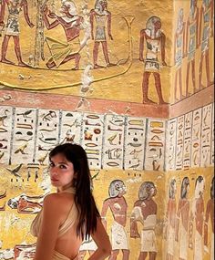 a woman standing in front of an egyptian wall with paintings on the walls behind her