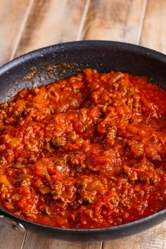 a skillet filled with meat and sauce on top of a wooden table next to a spoon