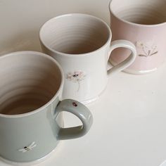 three coffee mugs sitting next to each other on a white counter top with flowers painted on them