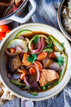 a bowl filled with meat and vegetables on top of a wooden table next to rice