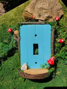 a blue fairy door with red mushrooms on the outside and grass growing around it in front of a rock