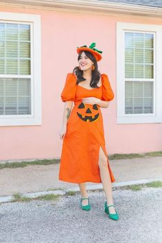 a woman in an orange dress and green shoes is standing near a pink house wearing a pumpkin costume