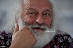 an older man with white hair and beard smiling at the camera while holding his finger to his chin