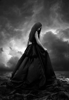 a woman in a long black dress standing on rocks with dark clouds above her and the ocean behind her