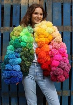 a woman standing in front of a wooden fence wearing a rainbow colored jacket with pom poms