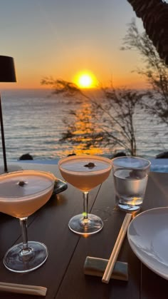 two martinis sitting on top of a wooden table next to the ocean at sunset