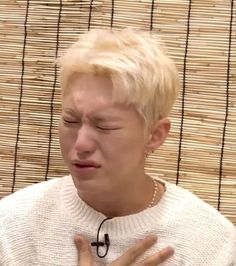 a man with his eyes closed sitting in front of a bamboo wall and wearing a white sweater