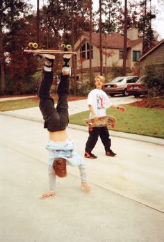 two young boys doing tricks on skateboards in front of a house, one holding up his hands and the other upside down