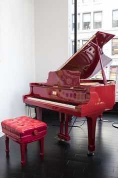 a red piano sitting on top of a hard wood floor next to a stool and window