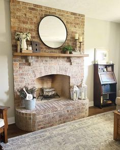 Candlesticks, potted plants, books, and a grand circular mirror decorate the top of this wooden mantel. Below the mantel, logs sit in the fire pit next to two white lanterns and a container filled with lavender stems...   Image: thecrowdedfarmhouse