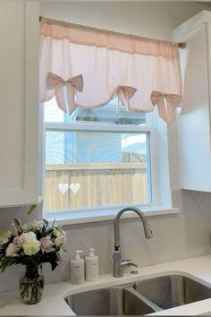 a kitchen sink under a window with pink bow valances on the windowsill and white cabinets