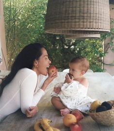 a woman is sitting on the floor with a baby eating an apple next to some bananas and apples