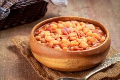 a wooden bowl filled with beans and bacon on top of a table next to a spoon