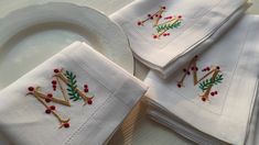 four white napkins with embroidered letters and holly on them, sitting on a plate