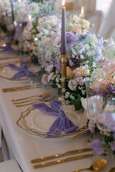 a long table is set with purple and white flowers, gold chargers and silverware