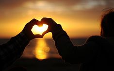 a person making a heart shape with their hands in front of the ocean at sunset