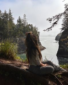 a woman sitting on the edge of a cliff looking out at the water and trees