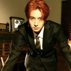 a man with red hair in a suit and tie posing for the camera while leaning on a table