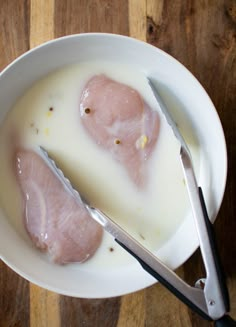 two pieces of raw chicken in a bowl with milk and utensils next to it