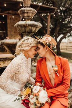 two brides sitting next to each other in front of a fountain wearing orange suits and hats