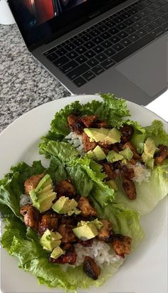 a white plate topped with lettuce covered in meat and avocado next to a laptop computer