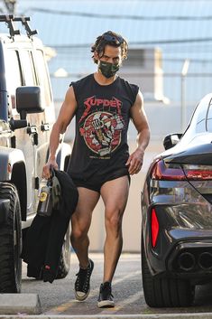 a man in black shirt and shorts walking down street next to cars with face mask on