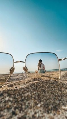 a woman sitting on top of a sandy beach next to the ocean reflected in her sunglasses