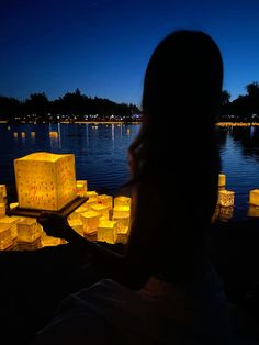 a woman holding a tray with paper lanterns floating in the water behind her at night