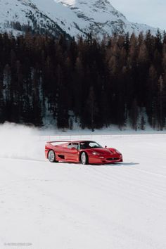 a red car driving down a snow covered road in front of some mountains and trees