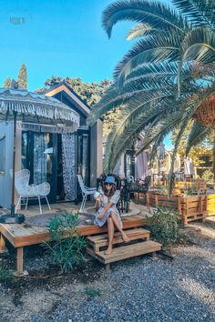 a woman sitting on a wooden bench in front of a small house with palm trees