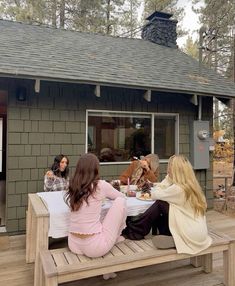 three women sitting at a picnic table with their dogs on the back porch in front of a cabin