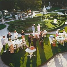 a group of people standing around a lush green yard with tables and chairs on it