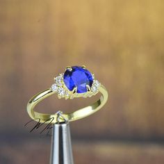 a gold ring with a blue stone surrounded by white diamonds on a wooden stand in front of a brown background