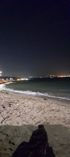 a person sitting on top of a sandy beach next to the ocean with lights in the background