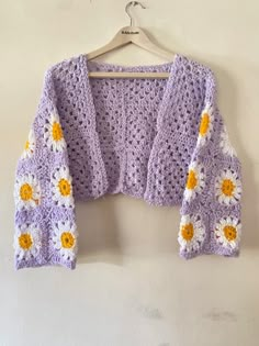 a purple crocheted sweater with yellow and white daisies on the shoulders hanging from a hanger