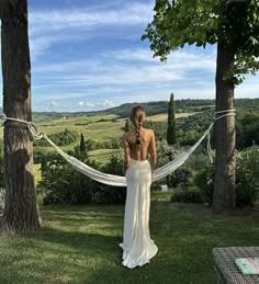 a woman in a white dress standing next to a tree with a hammock hanging from it's back