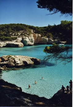 people are swimming in the blue water near some rocks and trees, while others stand on the shore