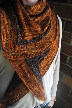 a woman wearing a knitted shawl in orange and black colors, standing next to a brick wall